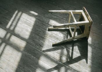 A worn wooden stool casting shadows on a sunlit wooden floor, creating a rustic and moody ambiance.