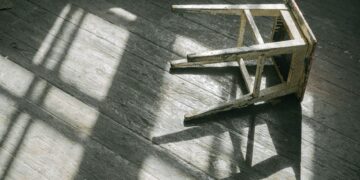 A worn wooden stool casting shadows on a sunlit wooden floor, creating a rustic and moody ambiance.