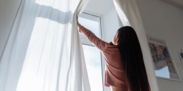 Woman opens white curtains in bright room, revealing a sunny day outside.