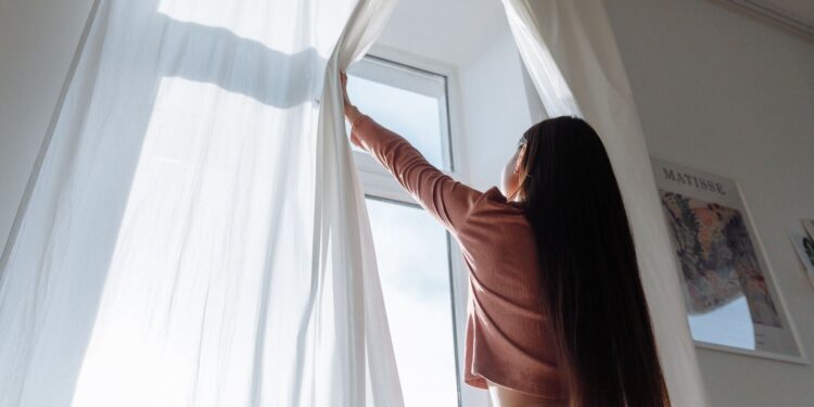 Woman opens white curtains in bright room, revealing a sunny day outside.