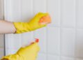 Crop unrecognizable woman in yellow rubber gloves cleaning white tiles with spray and soapy sponge