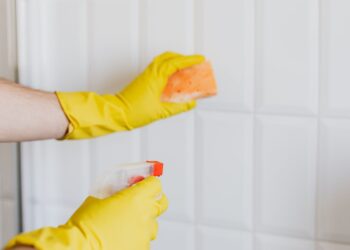 Crop unrecognizable woman in yellow rubber gloves cleaning white tiles with spray and soapy sponge