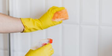 Crop unrecognizable woman in yellow rubber gloves cleaning white tiles with spray and soapy sponge