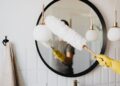 Crop anonymous female housewife in rubber gloves dusting modern lamp with soft brush against mirror while cleaning stylish bathroom
