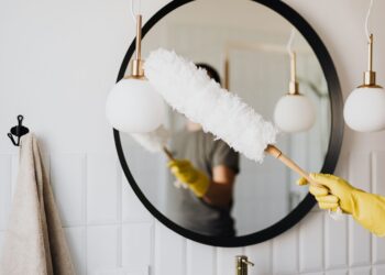 Crop anonymous female housewife in rubber gloves dusting modern lamp with soft brush against mirror while cleaning stylish bathroom