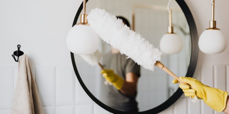 Crop anonymous female housewife in rubber gloves dusting modern lamp with soft brush against mirror while cleaning stylish bathroom