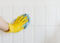 Crop anonymous housekeeper in yellow gloves cleaning white tiled wall using rag in bathroom