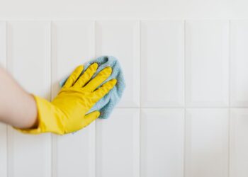 Crop anonymous housekeeper in yellow gloves cleaning white tiled wall using rag in bathroom