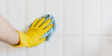 Crop anonymous housekeeper in yellow gloves cleaning white tiled wall using rag in bathroom