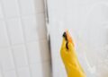 From above of crop anonymous person in yellow rubber protective glove washing shower cabin glass with sponge and detergent