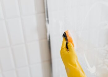From above of crop anonymous person in yellow rubber protective glove washing shower cabin glass with sponge and detergent