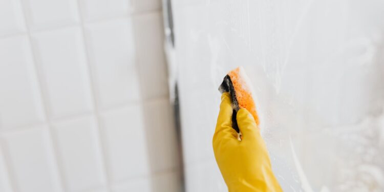 From above of crop anonymous person in yellow rubber protective glove washing shower cabin glass with sponge and detergent