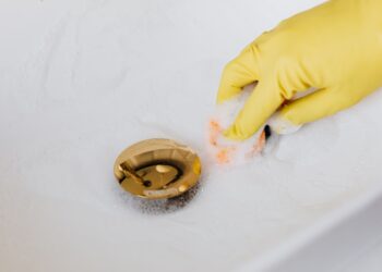 From above of crop anonymous person in yellow rubber protective glove using sponge and detergent for washing white sink in bathroom
