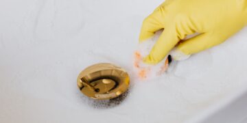 From above of crop anonymous person in yellow rubber protective glove using sponge and detergent for washing white sink in bathroom