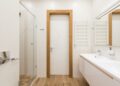 White door and furniture in modern bathroom with glass shower cabin and wooden elements reflecting in mirror