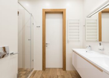 White door and furniture in modern bathroom with glass shower cabin and wooden elements reflecting in mirror
