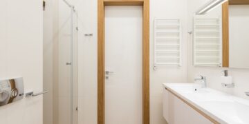 White door and furniture in modern bathroom with glass shower cabin and wooden elements reflecting in mirror