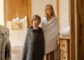 Two children stand wrapped in towels in a warmly lit, wooden bathroom with a washing machine.