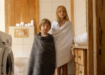 Two children stand wrapped in towels in a warmly lit, wooden bathroom with a washing machine.