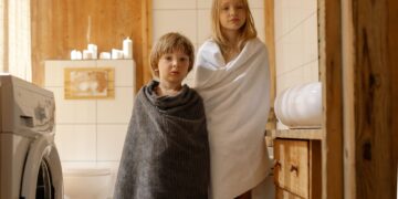 Two children stand wrapped in towels in a warmly lit, wooden bathroom with a washing machine.