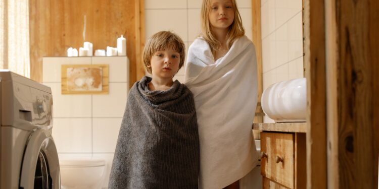 Two children stand wrapped in towels in a warmly lit, wooden bathroom with a washing machine.