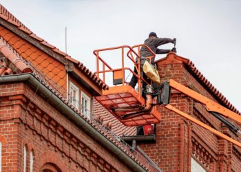 Roofer in a cherry picker conducting maintenance on a red brick building with a tiled roof.