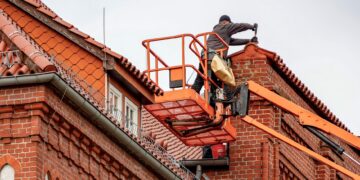 Roofer in a cherry picker conducting maintenance on a red brick building with a tiled roof.
