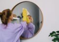 A woman wearing yellow gloves cleans a round wall mirror indoors, reflecting her image.