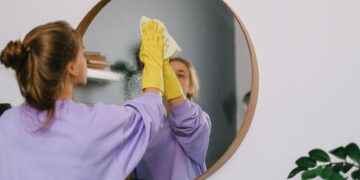 A woman wearing yellow gloves cleans a round wall mirror indoors, reflecting her image.