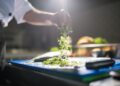 Chef's hand preparing chopped green vegetables on a cutting board in a kitchen.