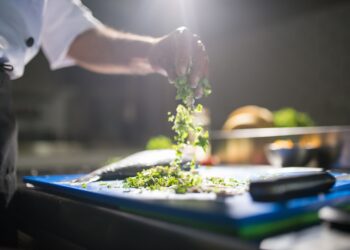 Chef's hand preparing chopped green vegetables on a cutting board in a kitchen.