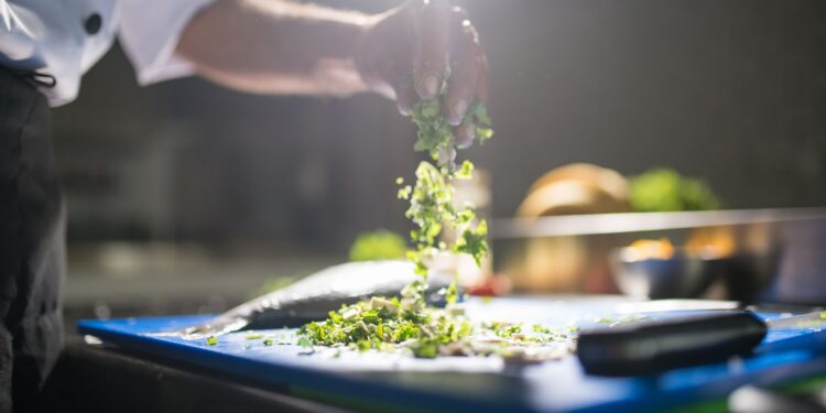 Chef's hand preparing chopped green vegetables on a cutting board in a kitchen.