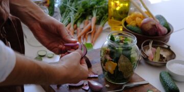 Close-up of hands preparing pickled vegetables in a jar with fresh carrots and cucumbers.