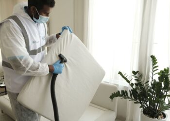 Man in protective gear vacuuming a mattress in a modern home interior.