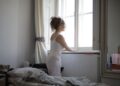 Back view of curly hair female wearing pajamas standing on bed and looking out of window after awakening in bedroom