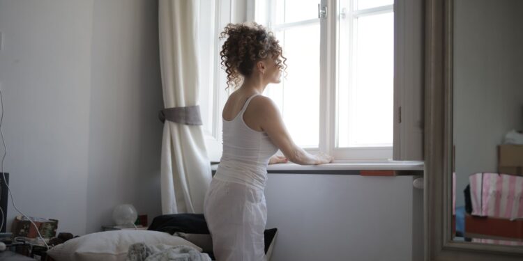 Back view of curly hair female wearing pajamas standing on bed and looking out of window after awakening in bedroom