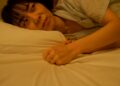 An Asian woman lying on a bed with a thoughtful and melancholic expression, gripping the pillow tightly.