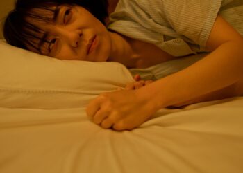 An Asian woman lying on a bed with a thoughtful and melancholic expression, gripping the pillow tightly.