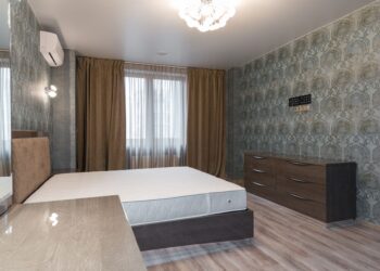 Interior of bedroom with air conditioning above bed placed near window with curtains and dresser