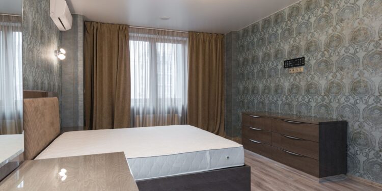 Interior of bedroom with air conditioning above bed placed near window with curtains and dresser