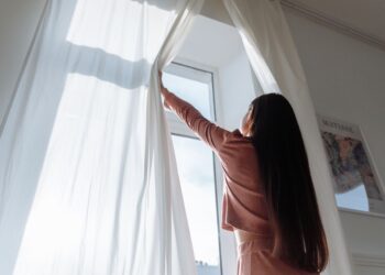 Woman opens white curtains in bright room, revealing a sunny day outside.