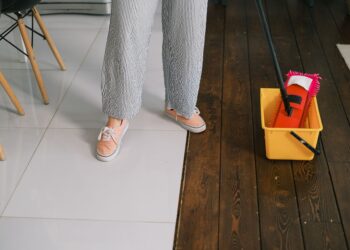 Crop anonymous woman in gumshoes and pants dipping mop into bucket with water on parquet in house