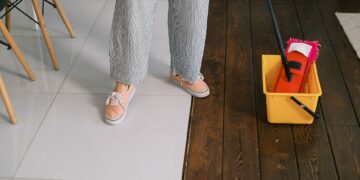 Crop anonymous woman in gumshoes and pants dipping mop into bucket with water on parquet in house