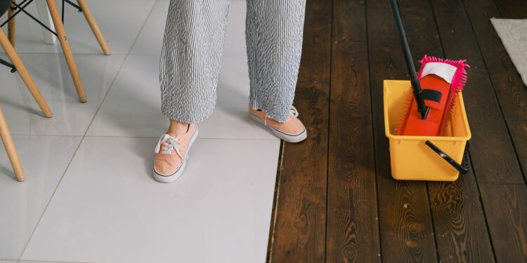 Crop anonymous woman in gumshoes and pants dipping mop into bucket with water on parquet in house