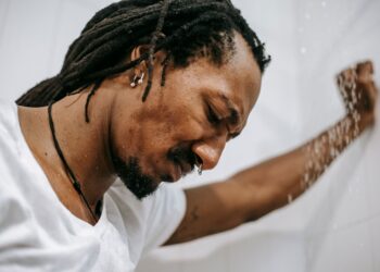 Crop irritated uncertain African American male in white short standing in shower cabin and frowning in dismay