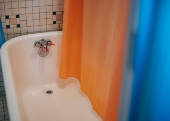 From above of clear white bath with steel faucet with hanging blue and orange curtain located in bathroom with tiled walls