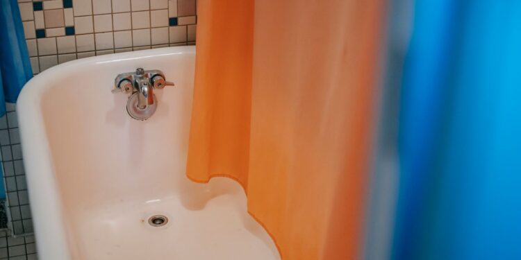 From above of clear white bath with steel faucet with hanging blue and orange curtain located in bathroom with tiled walls