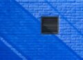 Close-up of a bright blue brick wall featuring a black ventilation grate and shadows.