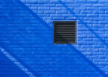 Close-up of a bright blue brick wall featuring a black ventilation grate and shadows.