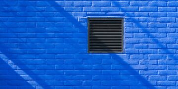 Close-up of a bright blue brick wall featuring a black ventilation grate and shadows.